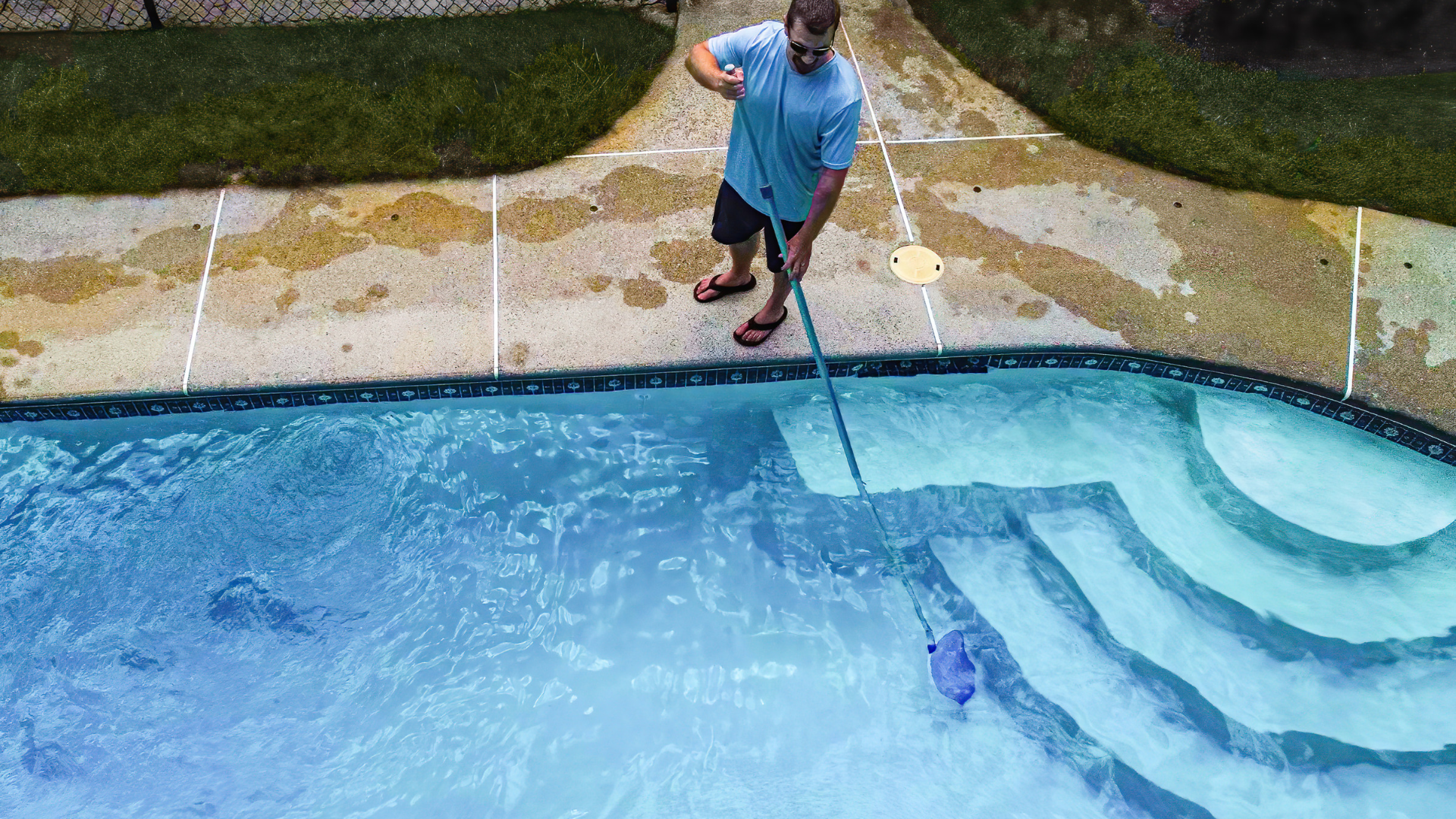 Pool cleaner using a long-handled net beside a blue swimming pool